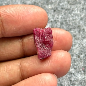 May include: A rough-cut, deep red ruby gemstone. The ruby has a textured surface and irregular shape, showcasing its natural, unpolished state. The gemstone is held between fingers, highlighting its size and color.