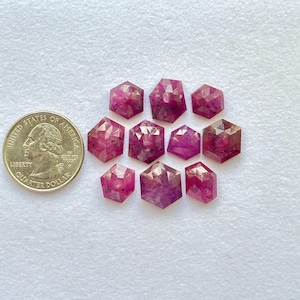 Op de afbeelding: Een verzameling gefacetteerde, zeshoekige robijn edelstenen in verschillende tinten roze en rood. Een Amerikaanse kwart dollar munt is inbegrepen ter vergelijking van de grootte. De edelstenen zijn gerangschikt op een wit oppervlak.