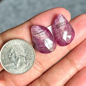 Op de afbeelding: Twee roze peervormige edelstenen met een facetgeslepen vorm. De edelstenen hebben een gestreept patroon en een zilveren glans.