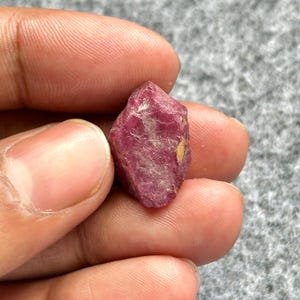 Peut inclure: Une pierre précieuse brute de rubis non taillée. Le rubis est d'un rouge profond et riche avec une surface légèrement inégale. La pierre est tenue entre deux doigts, mettant en valeur sa forme naturelle et brute. Le rubis a une petite tache jaune.