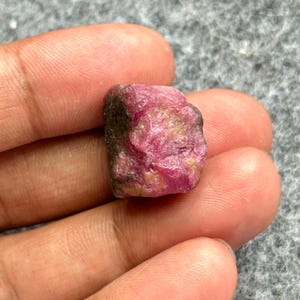 May include: A rough, uncut ruby gemstone. The ruby is a deep pinkish-red color with some darker areas and a slightly uneven surface. The gemstone is held in a hand, showing its natural, raw form.