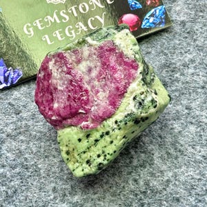 Puede incluir: Una gema en bruto con un vibrante cristal de rubí rosa incrustado en una matriz de zoisita verde, salpicada de negro. La imagen incluye una tarjeta con el texto "GEMSTONE LEGACY".