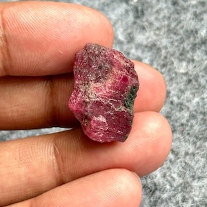 Peut inclure: Une pierre précieuse de rubis brute et non taillée, d'une teinte rouge profond, présentant des textures et des variations naturelles. Le rubis a une forme légèrement irrégulière et un aspect brut et terreux.