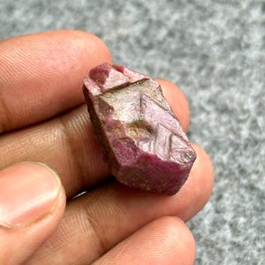 May include: A rough-cut ruby gemstone with a deep red hue and a slightly textured surface. The ruby has a natural, unpolished appearance, showcasing its raw form. The gemstone is held in a hand, highlighting its size and shape.