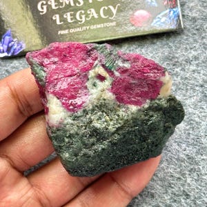 Puede incluir: Una gema en bruto con un vibrante color magenta, contrastada por depósitos minerales de color verde oscuro y blanco. La piedra se sostiene en una mano, con una tarjeta parcialmente visible en el fondo que dice "Gems Legacy, Fine Quality Gemstone."