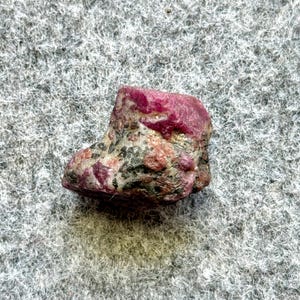May include: A rough, dark red ruby gemstone with a natural, irregular shape. The ruby has a dark red color with some black inclusions.