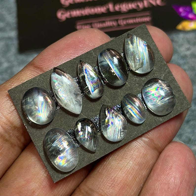 May include: A collection of ten clear, oval and teardrop-shaped gemstones with iridescent rainbow flashes. The gemstones are arranged on a dark gray rectangular card. The gemstones are held in a person's hand.