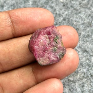 May include: A rough, uncut ruby gemstone with a deep red hue and some green inclusions. The natural stone has an irregular shape and a slightly textured surface. This raw ruby is a natural mineral specimen.