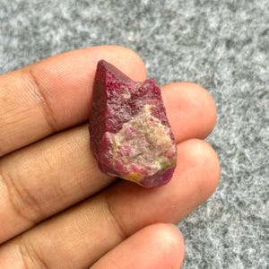 May include: A rough-cut, deep red ruby gemstone with a slightly uneven surface. The ruby has a natural, unpolished appearance, showcasing its raw, crystalline structure. The gemstone is held in a hand, highlighting its size and color.