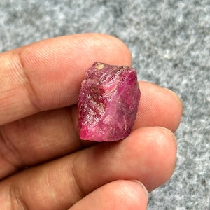 May include: A rough, uncut ruby gemstone. The ruby is a deep, vibrant red color with a slightly irregular shape. The gemstone is held in a hand, showcasing its natural texture and color.