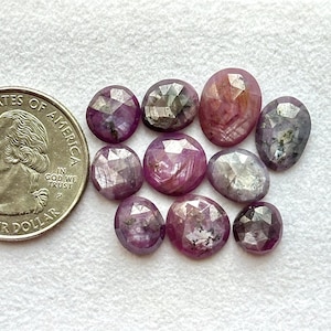 Pode incluir: Uma coleção de pedras preciosas facetadas, ovais e redondas, em tons de rosa e roxo, ao lado de uma moeda de um quarto de dólar americano para comparação de tamanho. As pedras preciosas têm uma aparência cristalina reflexiva e estão dispostas em uma superfície branca. Medidas em cm.