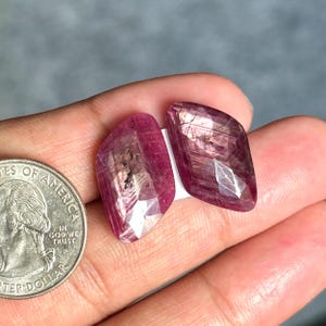 May include: Two faceted, kite-shaped pink rubies with visible internal inclusions and natural striations. The gemstones are positioned next to a United States quarter coin for scale, highlighting their translucent, deep magenta color and geometric cut.