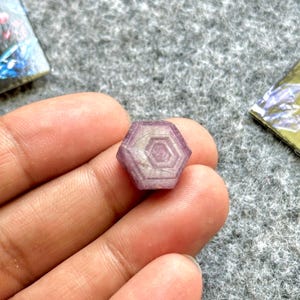 Puede incluir: Una pequeña gema hexagonal púrpura con un diseño concéntrico. La gema se sostiene en una mano, y el fondo es una superficie gris texturizada. Las facetas de la gema capturan la luz, resaltando su intrincado patrón.