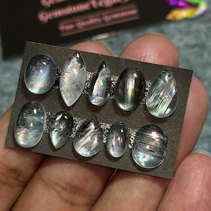 May include: A collection of ten iridescent gemstones in various shapes, including oval and teardrop, arranged on a dark brown card. The stones display a range of colors and internal patterns, with a shimmering effect. The gemstones are held by a hand.