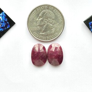 May include: Two oval-shaped, faceted, deep pink rubies positioned below a United States quarter coin for scale. The gemstones feature a polished, multi-faceted surface that reflects light, displaying a rich, saturated magenta-red color throughout.