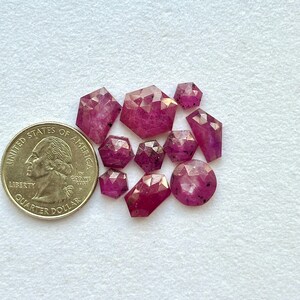 May include: A selection of faceted ruby gemstones in diverse shapes, including round, hexagonal, and rectangular. A U.S. quarter coin is placed beside the gemstones for size comparison. The rubies are a deep pinkish-red colour.