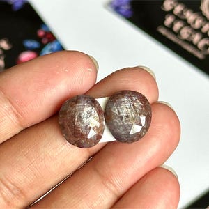 Pode incluir: São exibidas duas pedras preciosas ovais e facetadas com um tom castanho-avermelhado. As pedras são seguradas entre os dedos contra um fundo branco, com um cartão de visita parcialmente visível ao fundo. As pedras preciosas têm uma superfície polida e reflexiva.