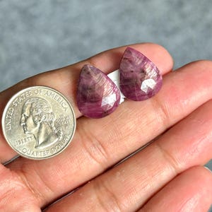 May include: Two faceted, teardrop-shaped ruby gemstones. The gems are a deep pinkish-red colour with internal inclusions. A U.S. quarter coin is next to the gemstones for size comparison. The gems are set on white paper.