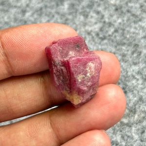 May include: A rough-cut, deep red ruby gemstone. The ruby has a natural, unpolished appearance with visible crystalline structures and a slightly uneven surface. The color is a rich, vibrant red with some lighter areas.