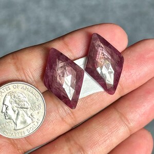 May include: Two faceted, diamond-shaped, deep magenta-coloured gemstones with visible internal inclusions and a polished surface, positioned next to a silver-coloured United States quarter coin for scale, showing the relative size of the stones.
