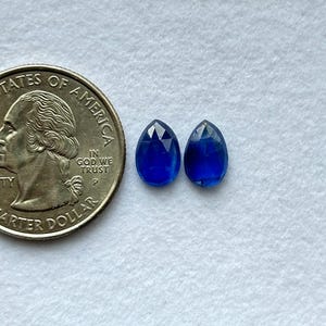 May include: Two faceted, teardrop-shaped, deep blue gemstones are displayed next to a U.S. quarter. The gemstones have a brilliant cut, reflecting light. The quarter provides a size reference, showing the gemstones' scale.
