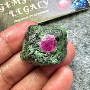 Puede incluir: Una gema en bruto con un centro de rubí rosa vibrante rodeado por una matriz verde. La piedra se sostiene en una mano, con el texto "Gems Legacy Fine Quality Gemstone" visible en el fondo. Es un espécimen natural sin pulir.