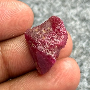 May include: A rough, uncut ruby gemstone with a deep red color. The ruby has a textured surface and irregular shape, showcasing its natural, raw form. The gemstone is held in a hand, highlighting its size and color.