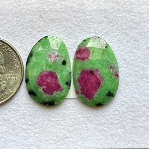 May include: Two oval-shaped, faceted zoisite gemstones with vibrant magenta ruby inclusions and small black spots. The stones are positioned next to a United States quarter coin for scale, displaying their polished surfaces and distinct color patterns.