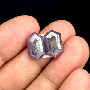 Pode incluir: São apresentados dois cristais hexagonais roxos com facetas refletoras. Os cristais são colocados sobre uma base branca e são segurados na mão de uma pessoa contra um fundo preto. Os cristais têm uma aparência natural e bruta.