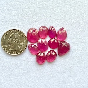 Puede incluir: Colección de gemas de rubí facetadas, de forma ovalada y de lágrima. Las gemas son de un color rojo rosado intenso y se muestran junto a una moneda para comparar el tamaño. Las gemas están sobre una superficie blanca.