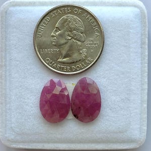 May include: Two oval-shaped, faceted pink rubies positioned below a United States quarter dollar coin. The gemstones exhibit a translucent quality with visible internal inclusions and a polished surface, shown for scale against the 0.955 inch coin.
