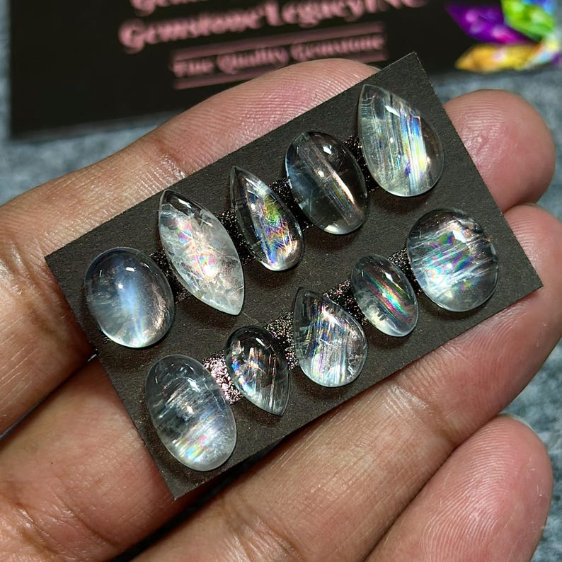 May include: A collection of clear, iridescent gemstones in various shapes, including oval and teardrop, arranged on a black card. The gemstones display a range of colors and internal patterns. The card is held in a hand.
