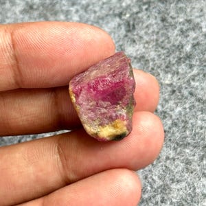 Peut inclure: Une pierre précieuse brute de couleur rose vif, avec des zones jaunes et vertes. La surface de la pierre est texturée, révélant son état naturel et non poli. La pierre est tenue dans une main.