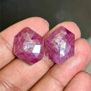 Pode incluir: Duas pedras preciosas hexagonais facetadas com um tom rosa profundo. As gemas têm múltiplas facetas que capturam a luz, criando um efeito brilhante. As pedras são seguradas em uma mão, mostrando seu tamanho e cor.