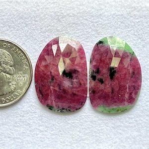 Puede incluir: Dos gemas ovaladas y facetadas con una moneda de veinticinco centavos para comparar el tamaño. Las gemas son principalmente de color rojo intenso con inclusiones negras y bordes verdes. Las facetas crean un efecto brillante.