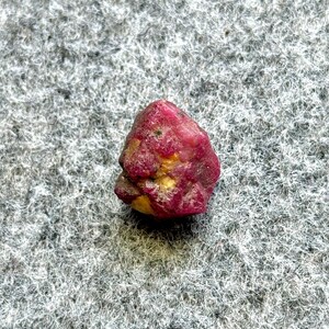 May include: A rough, uncut ruby gemstone. The ruby is a deep red color with some yellow and brown inclusions. The stone has a natural, irregular shape and is set against a gray, textured background.