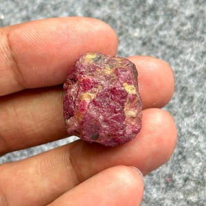 Könnte beinhalten: Ein roher, ungeschliffener Rubin-Edelstein. Der Rubin hat eine tiefrote Farbe mit helleren rosa und gelben Flecken. Der Stein hat eine natürliche, unregelmäßige Form und eine leicht raue Textur. Er wird zwischen zwei Fingern gehalten.