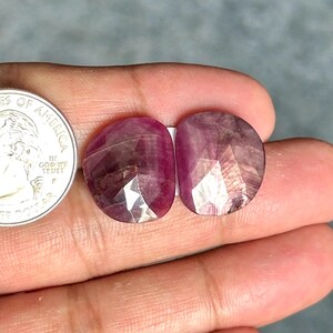 Puede incluir: Dos piedras preciosas facetadas en forma de lágrima de color magenta intenso, colocadas junto a una moneda de veinticinco centavos de dólar estadounidense para comparar el tamaño. Las piedras tienen una superficie pulida y multifacetada que refleja la luz.