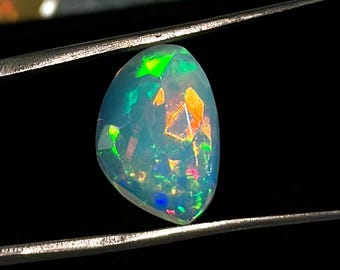 Könnte beinhalten: Ein polierter, tropfenförmiger Opal mit einer durchscheinenden, milchig-weißen Basis und lebhaften Blitzen in Neongrün, hellem Orange und tiefem Blau, der sicher von einer silberfarbenen Metallpinzette gehalten wird.