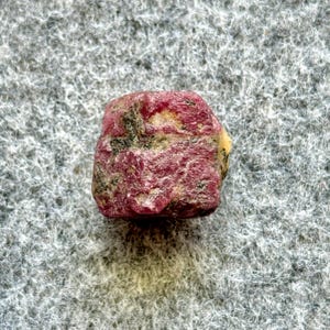 May include: A rough, red ruby gemstone with black and white inclusions. The ruby is a dark red color and has a slightly irregular shape.