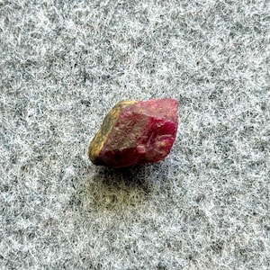 May include: A rough, uncut ruby gemstone. The ruby is a deep red color with areas of brown and gold. The stone has an irregular shape and a natural, unpolished appearance. The background is a textured gray surface.