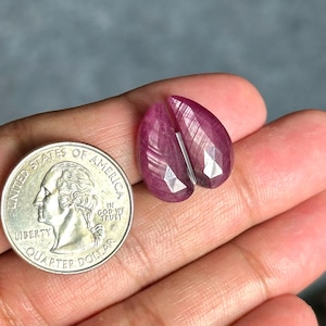 May include: Two faceted, teardrop-shaped, ruby gemstones with a quarter coin for size comparison. The rubies are a deep pinkish-red color and have a polished, reflective surface. The gemstones are set against a neutral background.