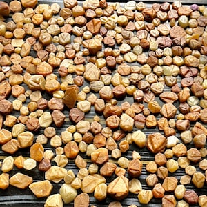 Puede incluir: Una gran colección de pequeñas piedras preciosas, ásperas, facetadas, de color marrón anaranjado. Las piedras preciosas están esparcidas sobre una superficie negra con un patrón rayado.