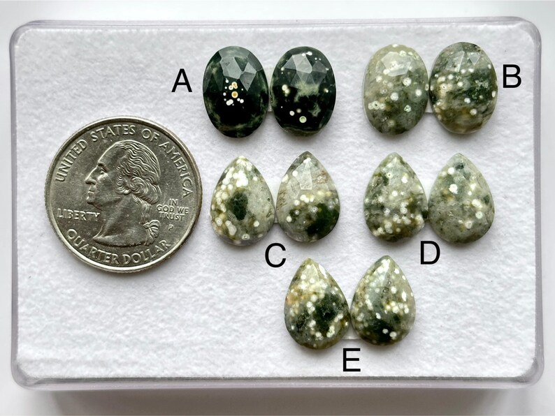 Op de afbeelding: Een verzameling edelstenen in verschillende vormen, waaronder ovaal en druppelvormig. De edelstenen zijn groen met witte spikkels. Een munt is inbegrepen ter vergelijking. De edelstenen zijn gelabeld met A, B, C, D en E. Afmetingen in centimeters.
