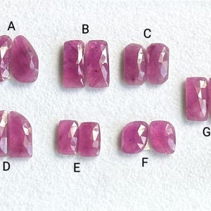 May include: Seven pairs of pink gemstone cabochons, labelled A to G, arranged in rows on a white surface.