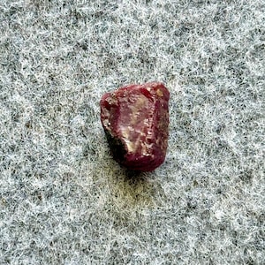 May include: A deep red ruby gemstone with a rough, natural shape. The ruby is sitting on a gray, textured surface.