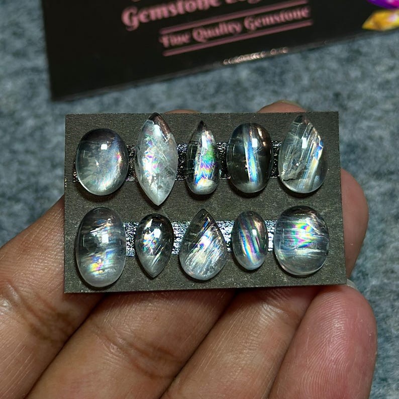 May include: A collection of ten iridescent gemstones in various shapes, including oval and teardrop, are displayed on a dark gray card. The gemstones exhibit a play of colors, with flashes of blue, green, and purple.