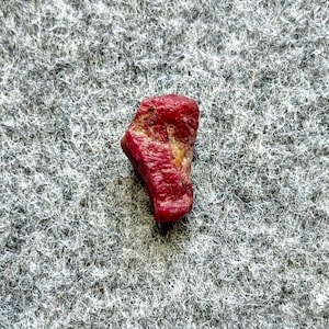 May include: A rough, uncut ruby gemstone with a deep red color and a slightly textured surface. The ruby has a natural, irregular shape and a small area of lighter, yellowish color.