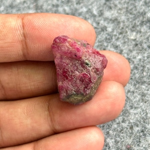 May include: A rough, uncut ruby gemstone with a deep red hue, held in a hand. The stone has a textured surface with areas of lighter pink and green. The gemstone is a natural, raw mineral.