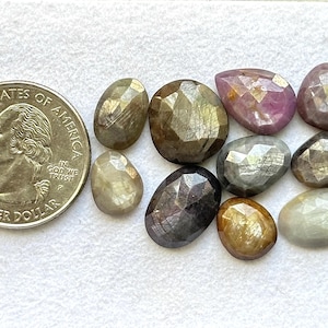 Pode incluir: Uma coleção de pedras preciosas facetadas em várias formas e cores, incluindo castanho, cinzento e rosa. Uma moeda de 25 centavos de dólar americano é colocada ao lado das pedras preciosas para comparação de tamanho. As pedras preciosas são exibidas em uma superfície branca.